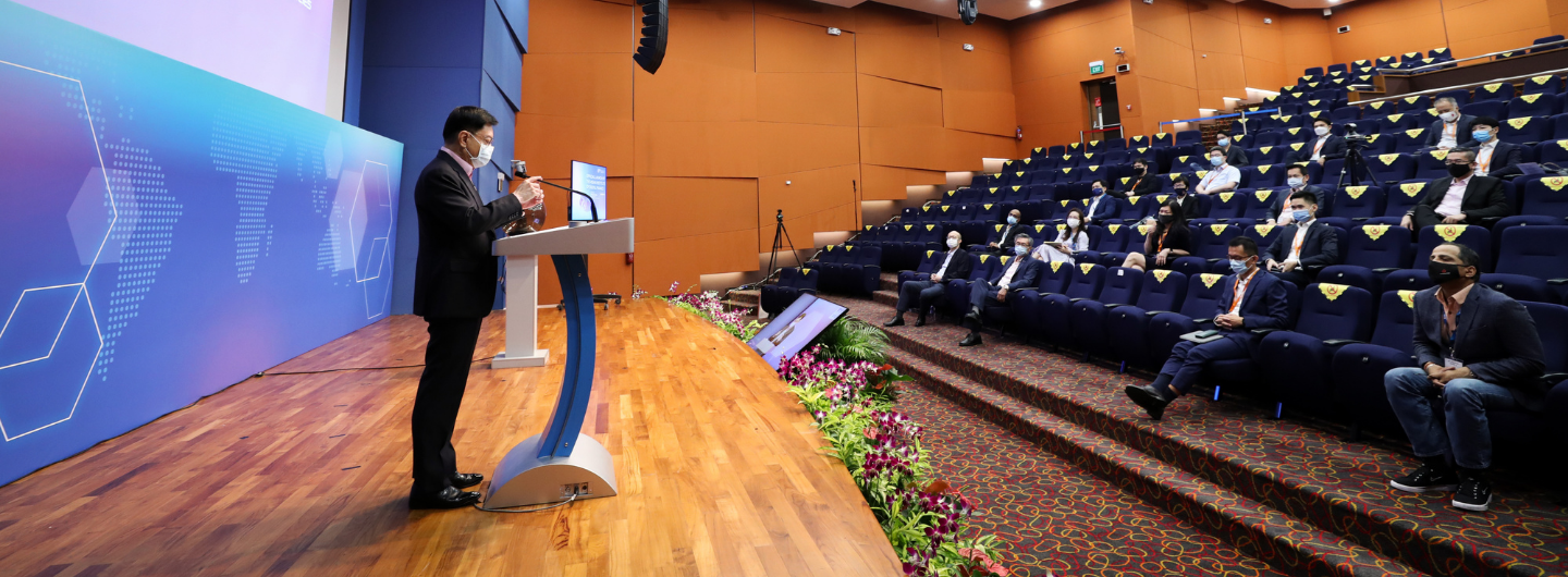 Speaker at podium on stage; audience seated wearing masks.